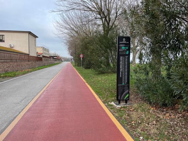 Ein roter Radweg verläuft parallel zu einer asphaltierten Straße mit einem digitalen Totem zur Zählung der Radfahrer auf der rechten Seite und einer von Bäumen gesäumten Einfahrt